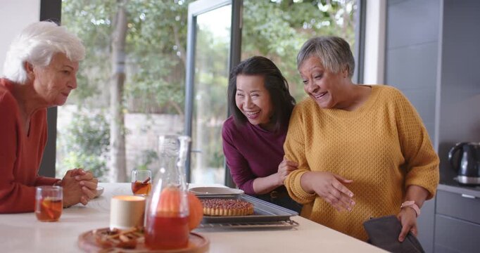Three women senior and middle-aged holding oven mitts, placing tart on tray in kitchen, admiring