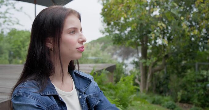 Woman sitting arms folded on deck beside parasol, in denim jacket, turning toward pond then camera