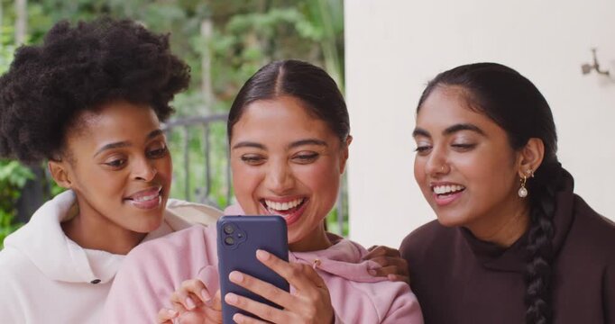 Female friends in hoodies leaning as center woman tapping blue phone on porch laughing at clip