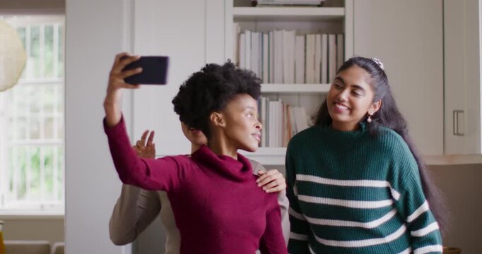 Diverse female friends with Black woman raising smartphone, adjusting poses to fit faces in kitchen