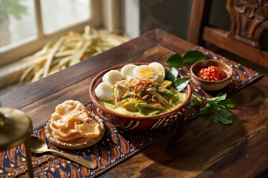Indonesian sayur lodeh vegetable coconut milk soup bowl served with rice cakes crackers and sambal on rustic wooden table with traditional batik fabric.