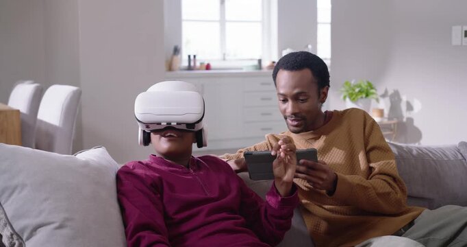 African American dad handing remote and tapping tablet guiding teen son wearing VR headset on couch
