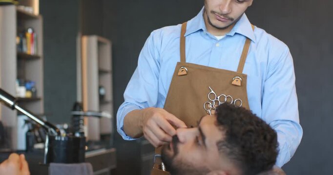 Male barber and male client reclining, barber grooming moustache with trimmer at barbershop
