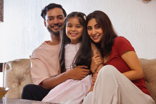 Young couple with their daughter looking at camera while sitting together on sofa at home