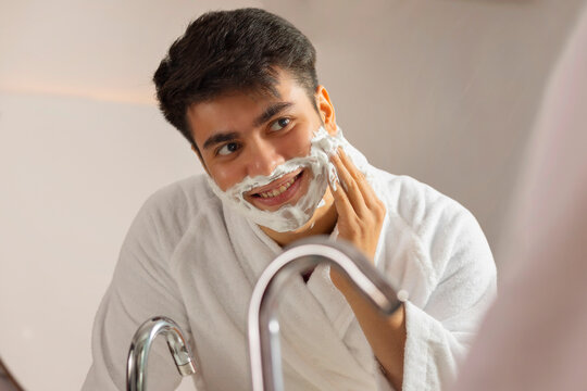 Close-up portrait of young man applying shaving cream to his face in a bathroom at home