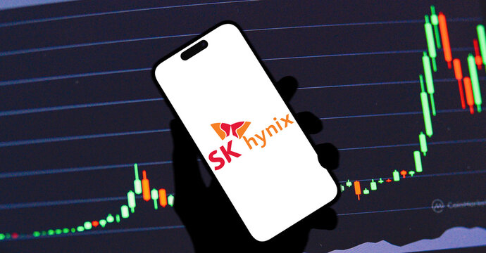 SK Hynix Logo Displayed on Tilted Smartphone Screen Held in Silhouette Hand Against Stock Market Candlestick Chart Background