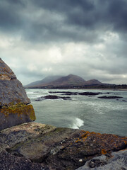 Fototapeta premium Stone pier stretches into ocean under dark storm clouds. Water crashes against rocks at shore. Famous Croagh Patrick Mountain rises in distance behind calm water, County Mayo, Ireland. Irish landscape