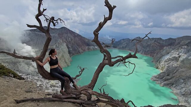 Wide shot of a young woman sitting on a bare dead tree at the edge of Mount Ijen volcanic crater looking at the turquoise acid lake. Travel fitness lifestyle.