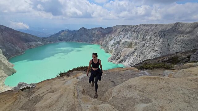 Wide shot of a sporty young woman hiking along the rugged path of Mount Ijen volcanic crater with a scenic turquoise acid lake background. Active lifestyle.