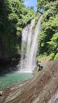 Vertical static shot of Gitgit Waterfall cascading into a turquoise natural pool on Bali island, surrounded by lush green jungle foliage. Nature stock.