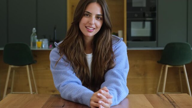 A beautiful young woman with long, dark hair smiles genuinely at the camera from her modern kitchen table. Her warm, friendly expression conveys happiness and positive energy, making for an inviting v