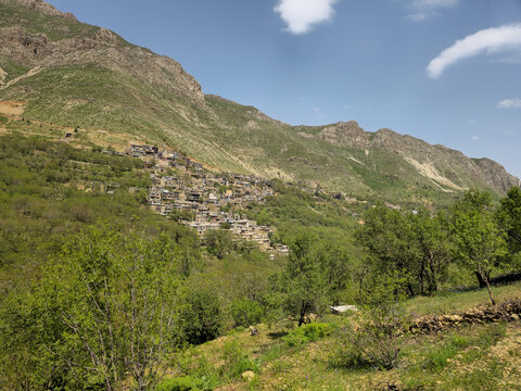 Traditional stone houses form a mountain village on a lush green hillside beneath rugged peaks, surrounded by spring orchards under a clear blue sky.
📍 Kamalla village, Hawraman Takht, uraman takht 