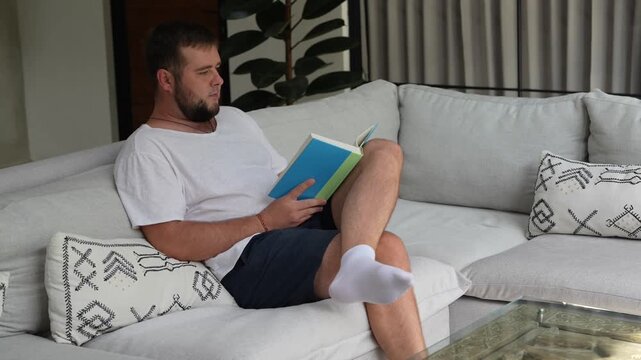 Relaxed man reading a book while sitting comfortably on a sofa at home. Quiet leisure time, reading habit, home comfort and peaceful indoor lifestyle.