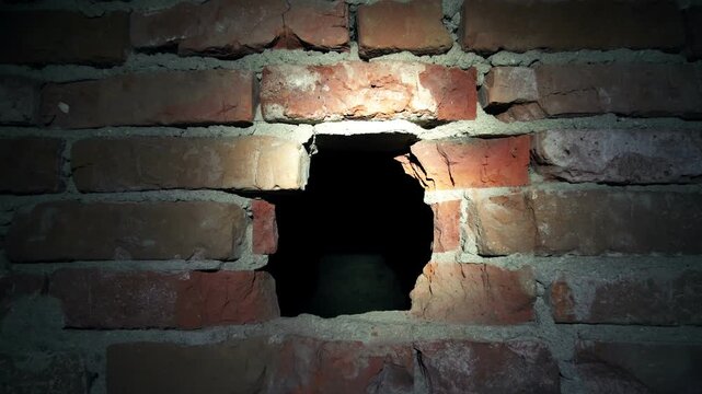 Flashlight beams sweep across a rough brick wall with a small broken opening leading into a dark interior cavity, creating a moody abandoned urban scene