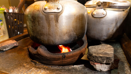Fototapeta premium Two old, metallic kettles are being heated on a traditional clay stove with glowing embers, showcasing a rustic cooking scene.