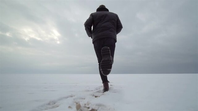 Rear view of a lone person walking across a vast snowy plain, leaving footprints and then pausing beneath a heavy overcast winter sky