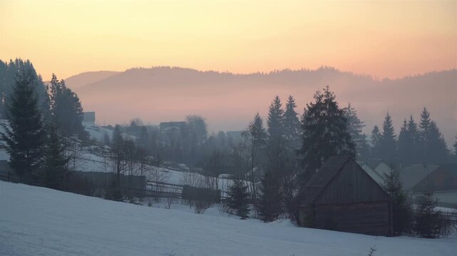 Misty winter sunrise over a snowy rural hillside with wooden barns, scattered houses and dark conifer trees silhouetted against layered forested hills