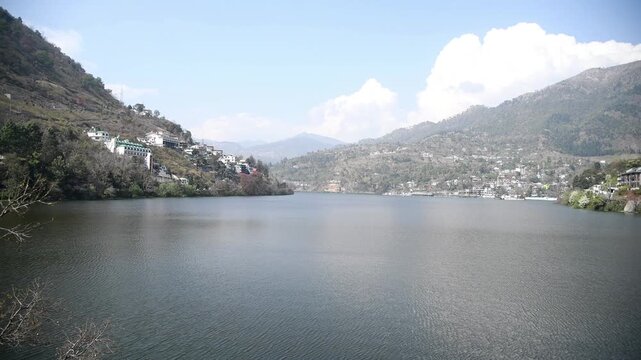 A wide panoramic shot of the beautiful Bhimtal Lake in Uttarakhand, India. The calm blue water of the lake is surrounded by lush green and rocky hills dotted with local houses, hotels and resorts. 