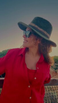 A happy woman enjoys the golden hour outdoors, wearing a stylish straw hat, a vibrant red shirt, and cool sunglasses. She smiles warmly, soaking in the serene evening light and peaceful atmosphere, em