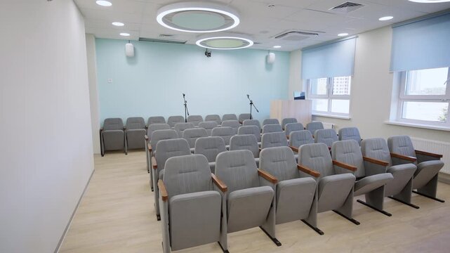 Spacious assembly hall contains rows of cushioned chairs with gray fabric upholstery. Microphones and stage prepared for conferences