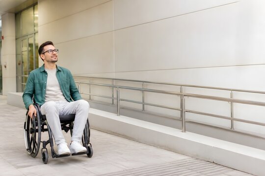 Professional young man in a wheelchair smiling and looking away in a modern office building exterior with copy space for commercial stock photography, depicting inclusivity and workplace mobility.