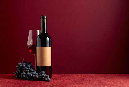 Bottle and glass of red wine with blue grapes on a red background.