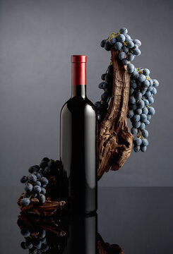 Bottle of red wine with a weathered snag and blue grapes on a black reflective background.