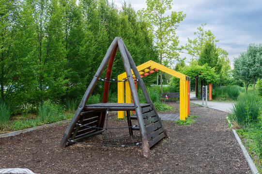A triangular timber climbing frame with ropes and platform stands beside yellow and orange monkey bars in a green public park. The empty recreation area offers equipment for climbing, balance, agility