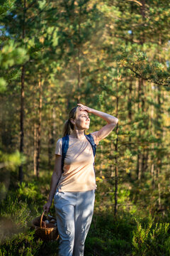 Woman forager taking break to enjoy nature and sunlight while harvesting mushrooms in forest, calm female observing surroundings standing in woods looking away, recreational activity and relaxation.