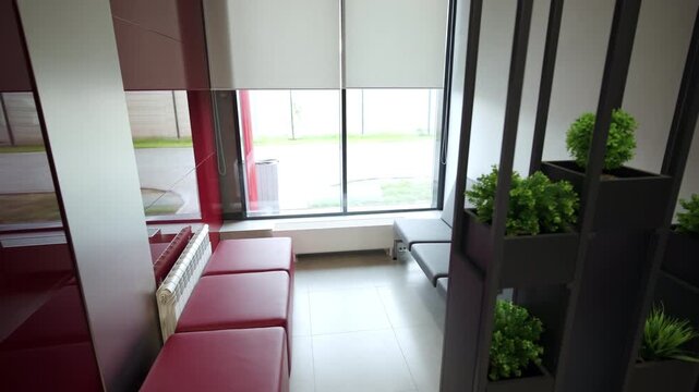 Cozy waiting zone features minimalist chairs and rows of red square ottomans. Large window with blinds fills small space with daylight