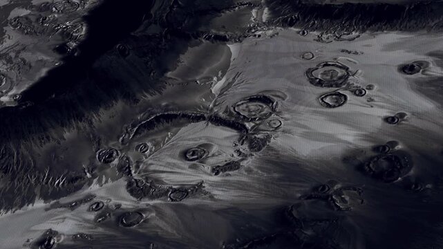The view shows multiple volcanic craters on the surface of a distant planet. The landscape appears dark with circular formations. This scene captures the surface details in space.