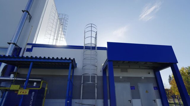 Modern industrial building facade with bright blue metal structures. Vertical safety ladders and steel pipe stretch toward clear sky