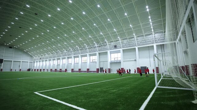 Football players in uniforms train on artificial turf in huge indoor arena. Massive arched ceiling with spotlights illuminates field