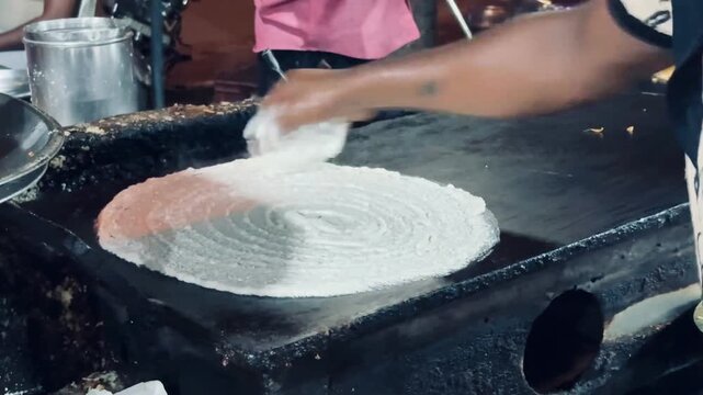 Close-up of a chef skillfully preparing a crispy dosa by spreading fermented rice batter on a hot tawa. The image captures the traditional cooking process of one of India's most iconic dishes.