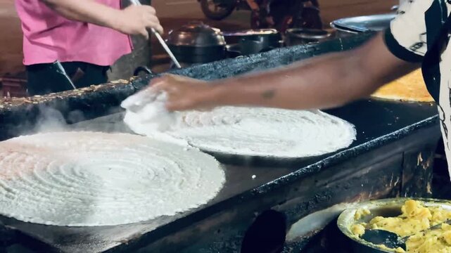 A skilled street food vendor spreads fresh dosa batter evenly on a hot iron griddle, showcasing the traditional preparation of South Indian cuisine.