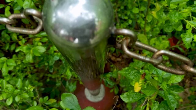 Close up shot of stainless steel bollard post top with linked metal chain among green leaves