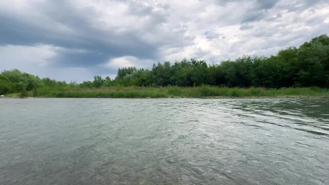 A calm river flows past green grassy banks under a cloudy sky. Dense summer trees grow along the opposite shoreline in daylight as water surface ripples gently through this natural landscape.