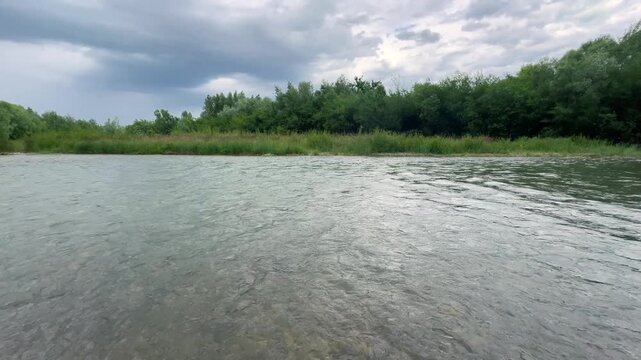 Scenic view of a wide river flowing past a lush green forest under a cloudy sky. Camera captures natural movement of water ripples with a dense line of trees on the opposite countryside bank.