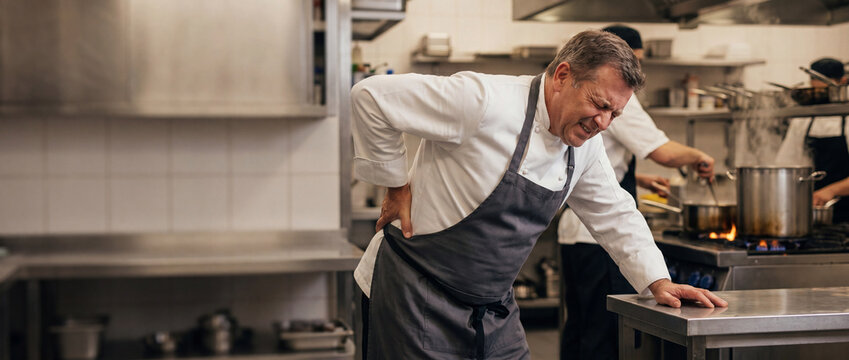 Chef suffering from back pain in kitchen, grimacing in discomfort while leaning on a stainless steel counter during a busy restaurant shift, ideal for medical, occupational health