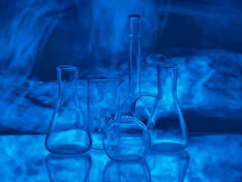 Empty glass laboratory flasks and retorts in dark blue neon light with mysterious smoke for scientific research, biotechnology, medical innovation and pharmacy background
