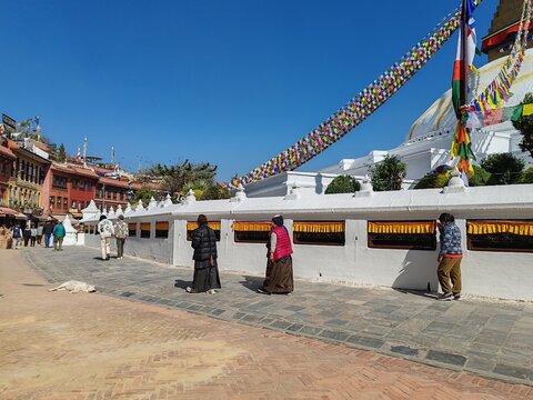 nepal religious site with white stupa, colorful prayer flags, people walking on a cobblestone path under clear blue sky