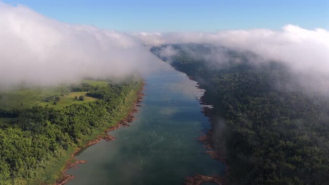 Aerial view of a wide river cutting through dense morning clouds along a forested border landscape.