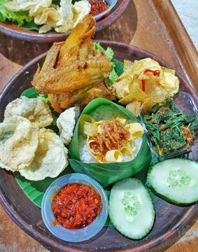 Authentic Indonesian fried chicken served with steamed rice wrapped in banana leaf, sambal, vegetables, cucumber and crackers. Traditional Southeast Asian comfort food on rustic wooden table.