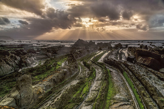 Powerful sunbeams breaking through dark storm clouds over mossy flysch rock formations at Barrika beach coast in Basque Country Spain