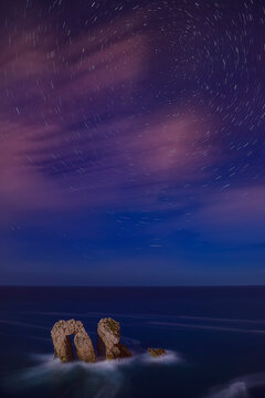 Stunning long exposure astrophotography showing circular star trails in the night sky over sea stack rock arches of Los Urros in Cantabria Spain