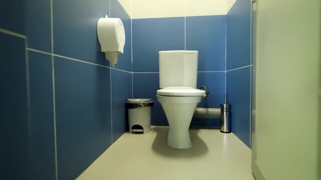 Ceramic toilet stands in clean public restroom cubicle with tiled walls. Small waste bin and paper dispenser complete functional space