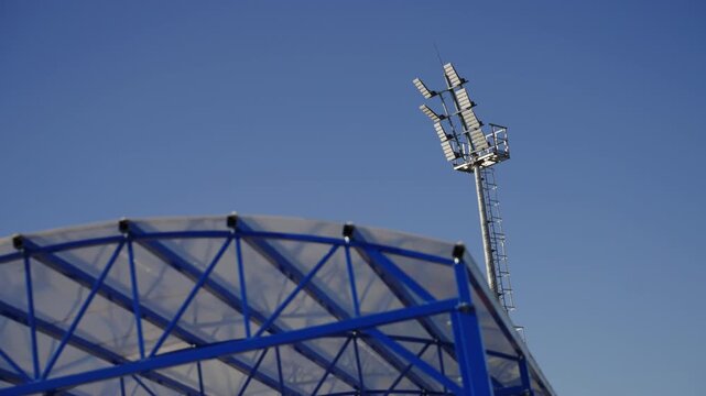 Stadium floodlight mast stands tall above canopy structure. Clean facility looks ready for evening training and matches under clear sky
