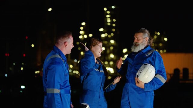 Professional industrial team of diverse engineers in blue uniform celebrating success with high fives at night at an oil refinery plant. Achievement, teamwork, and energy industry concept in a factory