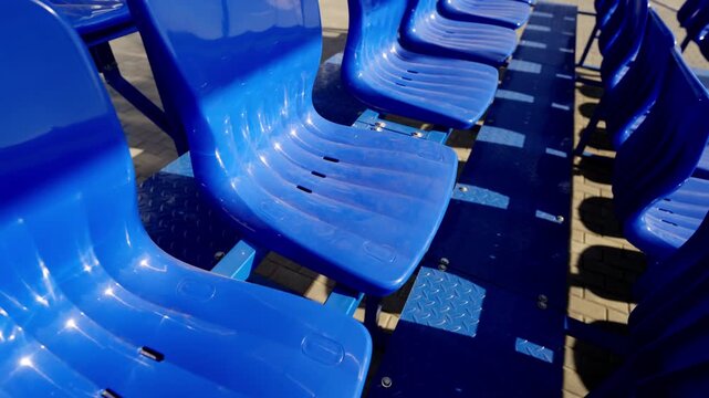 Plastic seats stand empty on outdoor metal tribune structure. Sunlight creates sharp shadows and reflections on glossy clean surfaces