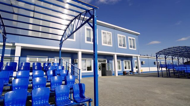 Rows of glossy spectator chairs form covered stands at regional public sports venue. Sunny afternoon weather illuminates paved ground